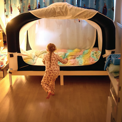 A child in leopard-print pajamas runs toward a large white-and-black canopy bed with pastel blankets in a cozy kid's room to the side of a bright, warmly lit space.