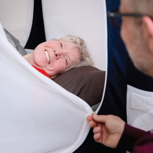 Smiling elderly woman lying in a white mesh medical cocoon being zipped shut by a caregiver.