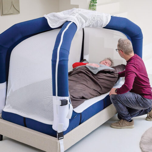 Caregiver kneels beside an elderly person lying in a large padded blue-and-white bed or therapy pod, assisting with positioning under a brown blanket.