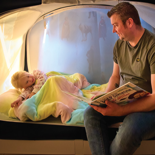 Man sitting on a bed inside a pop-up tent, reading a newspaper to a young child tucked in pastel blankets. The cozy scene suggests a bedtime story moment.