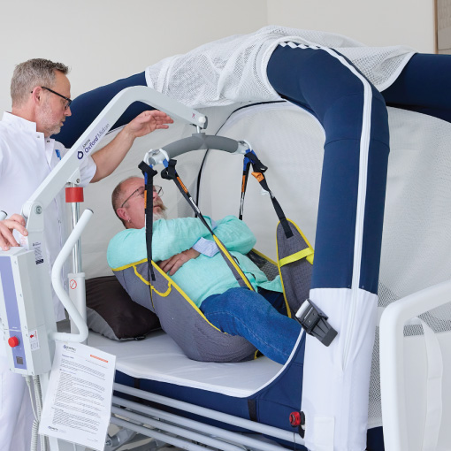Healthcare professional helps a patient into a blue-and-white medical rehabilitation chair with a harness in a clinic setting.