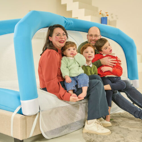 Family of four (mother, father, and two young children) sitting together on a blue padded indoor play structure, smiling at the camera.