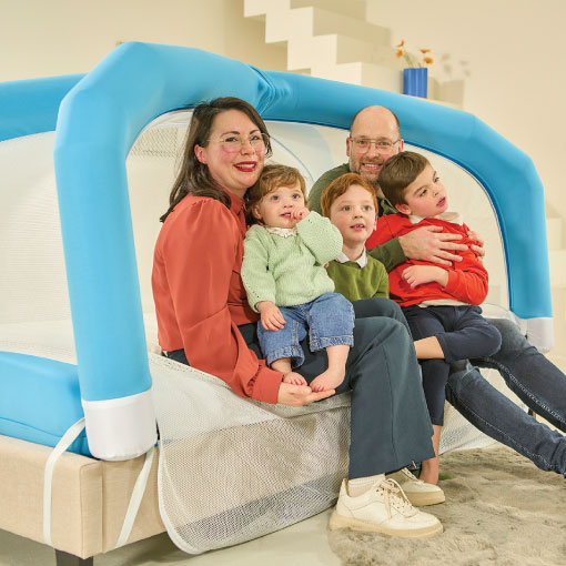 Family of four (mother, father, and two young children) sitting together on a blue padded indoor play structure, smiling at the camera.