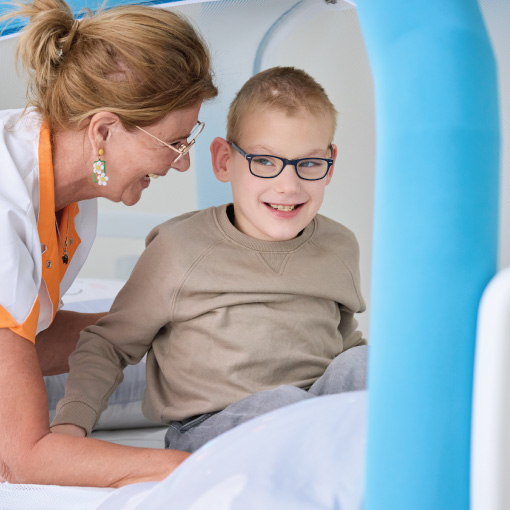 Smiling boy with glasses sits on a medical exam bed as a female clinician leans in to talk with him in a bright clinical setting.