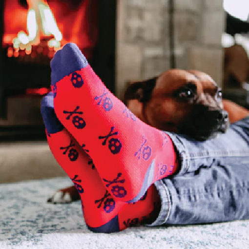Person lounging on the floor with feet in pink socks featuring black skull patterns, beside a dog by a lit fireplace.