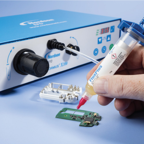 Technician applies flux from a syringe to a small green circuit board while inspecting a dismantled device near a blue lab instrument with a display panel and cables.