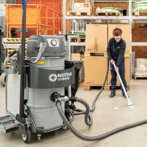 Industrial vacuum cleaner labeled Nilfisk VHB436 in a warehouse, hoses connected, worker sweeping the floor with a broom nearby.