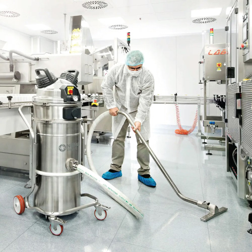 Person in a cleanroom suit and blue shoe covers operates a floor-cleaning machine in a bright pharmaceutical processing room